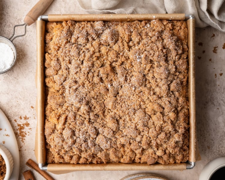 Baked cinnamon crumb cake in the pan with golden, textured streusel topping; next frame shows the cake dusted lightly with powdered sugar.