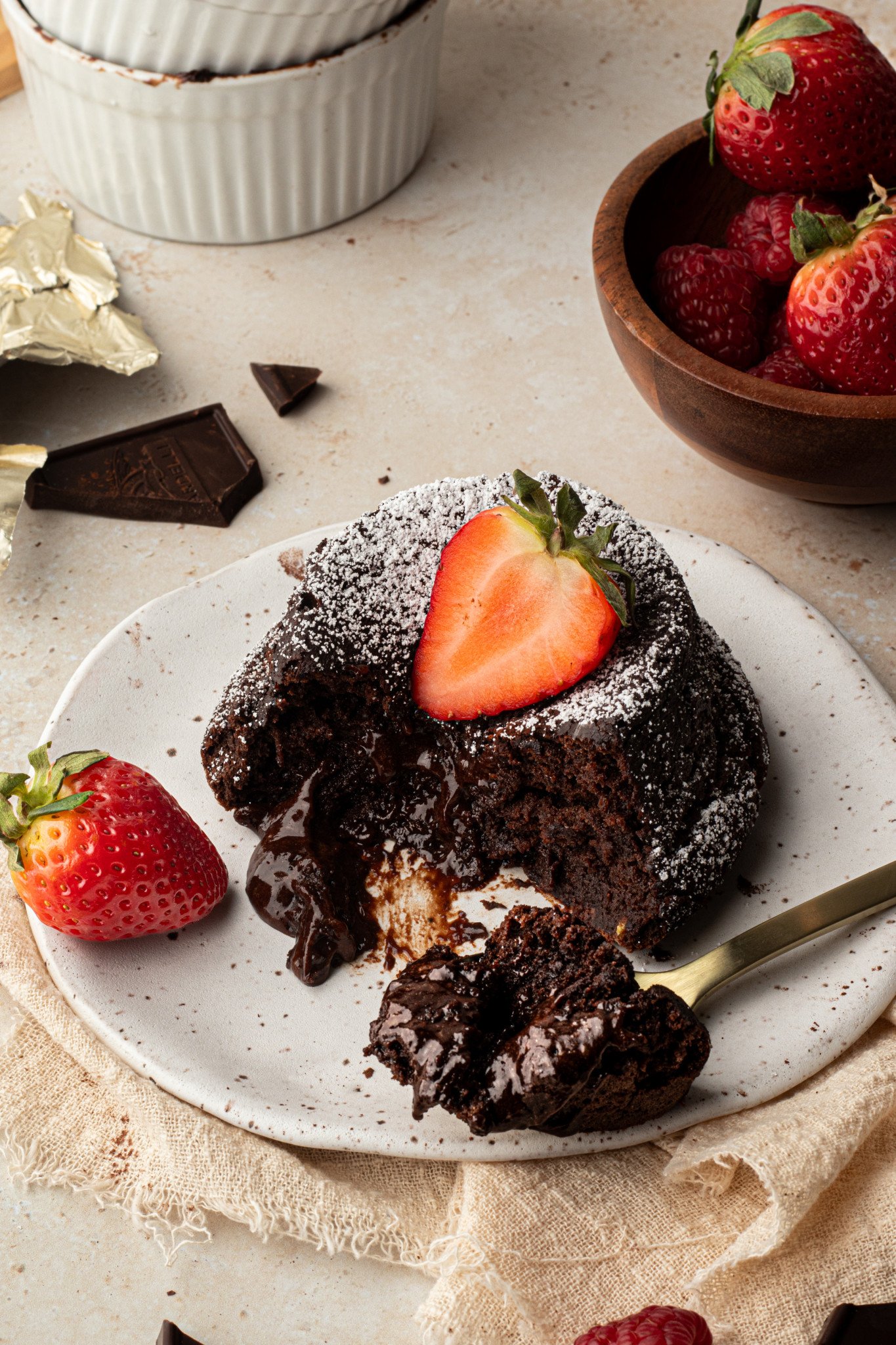 chocolate molten cake on a plate with a fork taking a bite and strawberries and powdered sugar for garnish. 