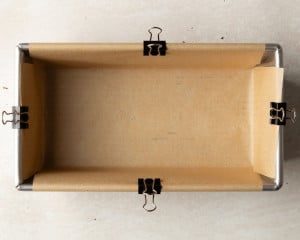 A metal loaf pan lined with parchment paper secured neatly with black binder clips along the edges, ready for brownie batter.