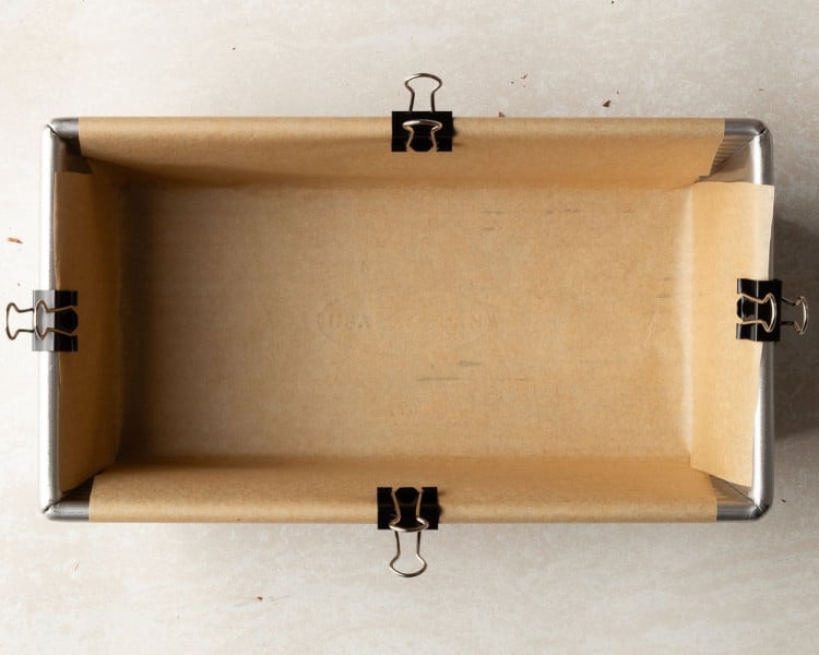 A metal loaf pan lined with parchment paper secured neatly with black binder clips along the edges, ready for brownie batter.