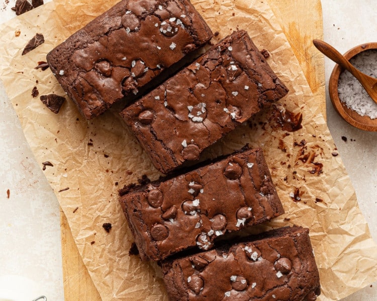 A close-up view of sliced fudgy brownies on parchment paper with scattered sea salt flakes and melty chocolate chips, set on a wooden cutting board.