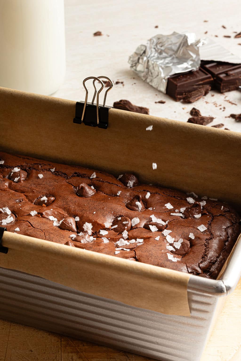 A close-up side view of a loaf pan filled with baked brownies. The brownies are topped with chocolate chips and flaky sea salt, and bits of broken chocolate and foil sit in the blurred background.
