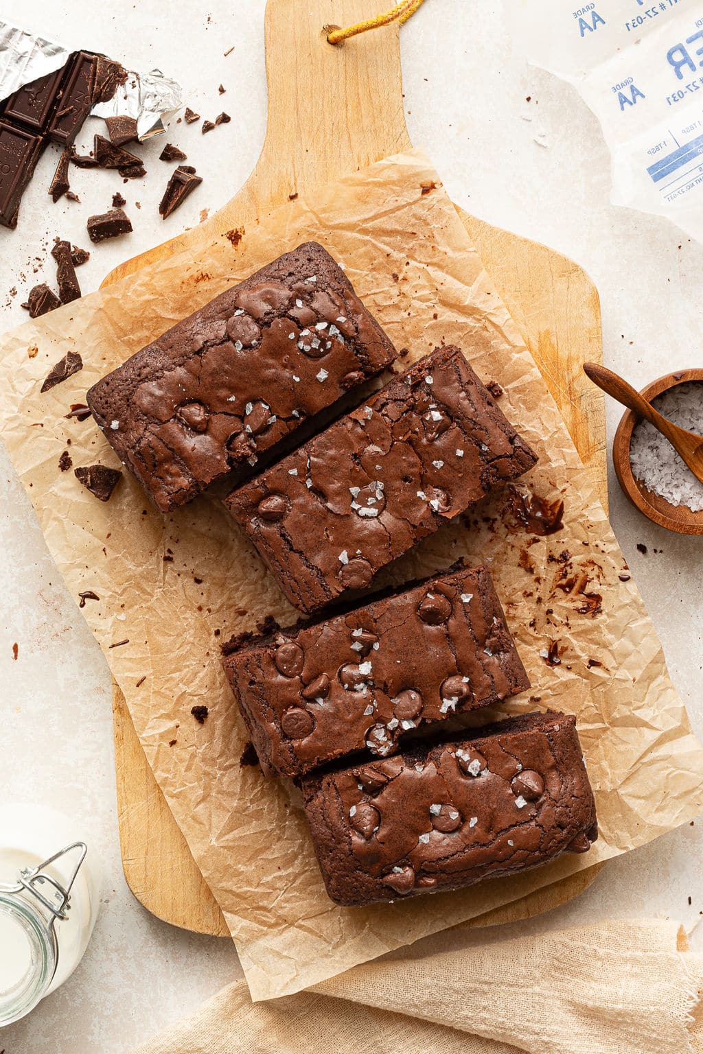 Four sliced brownies arranged on parchment paper, showing their crinkly tops with chocolate chips and sea salt. Surrounding details include broken chocolate pieces and a wooden cutting board.