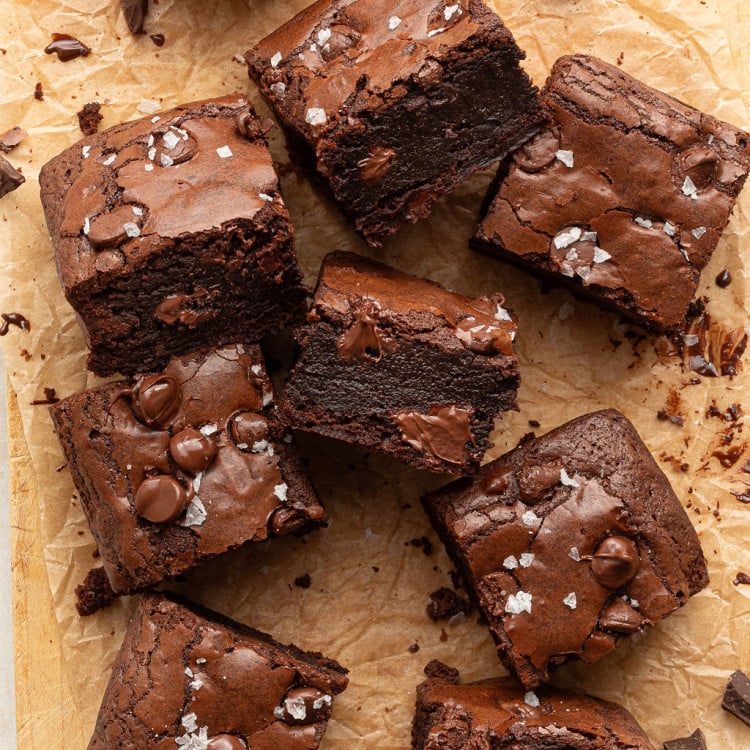 Several fudgy brownie squares, some stacked and one with a bite taken out, are arranged on parchment. The tops are sprinkled with flaky sea salt and melty chocolate chips.