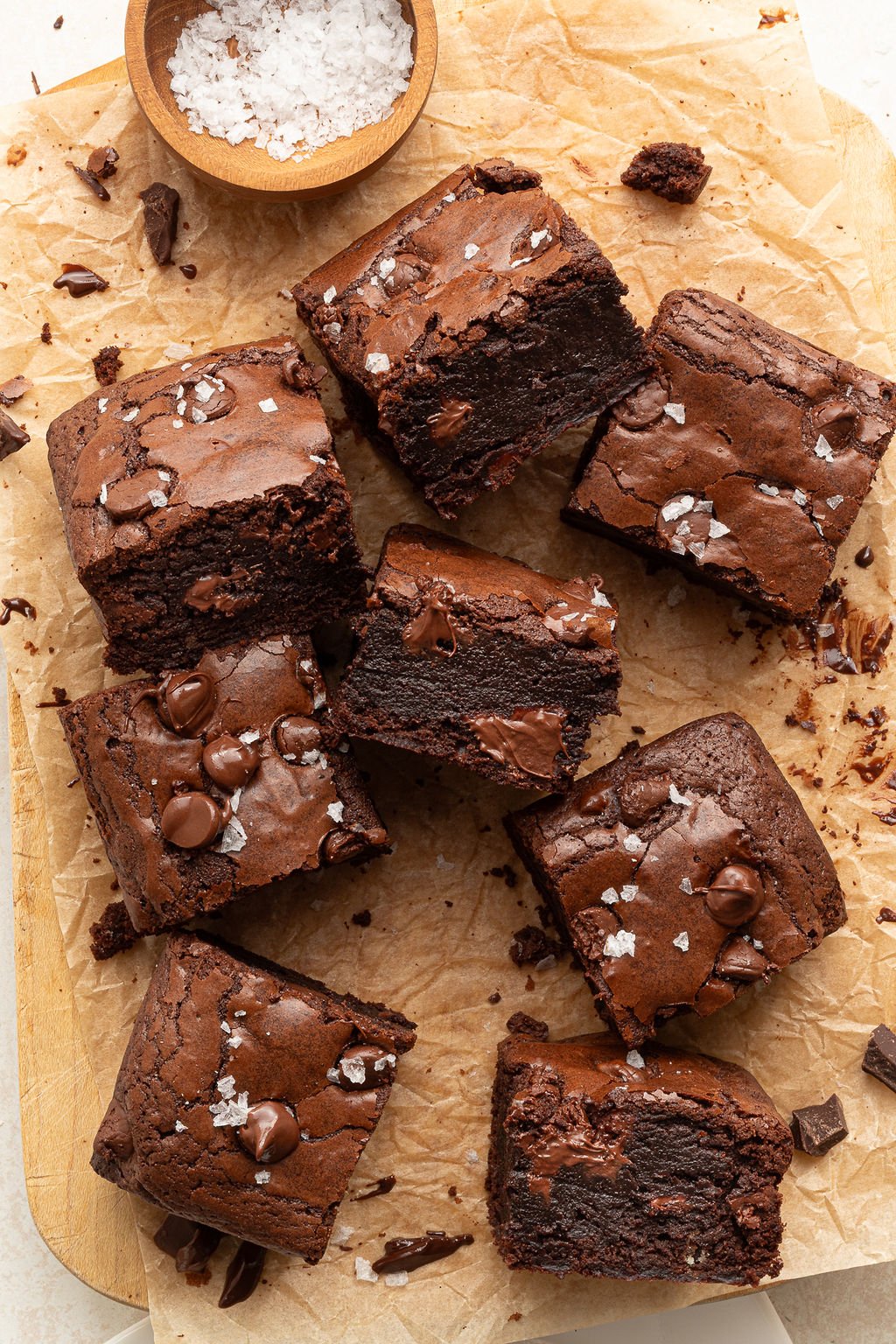 Several fudgy brownie squares, some stacked and one with a bite taken out, are arranged on parchment. The tops are sprinkled with flaky sea salt and melty chocolate chips.