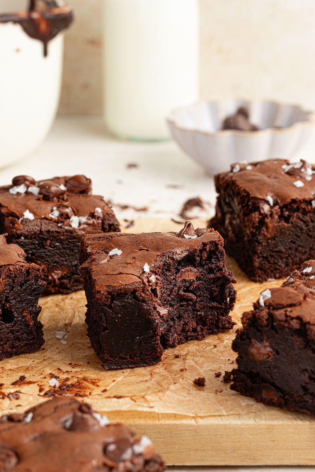 Side view of sliced brownies on a wooden surface. The center brownie shows a rich, gooey interior. A milk bottle and a bowl of chocolate chips appear softly blurred in the background.