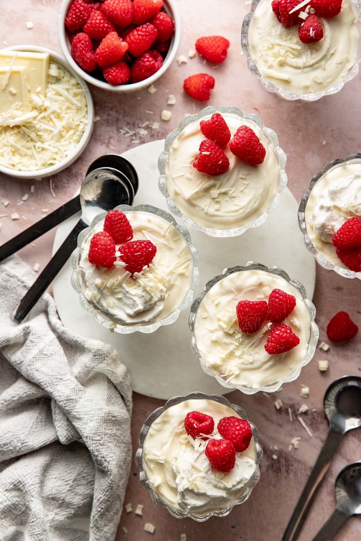 Overhead photo of several glass dessert cups filled with creamy white chocolate mousse, topped with fresh raspberries, whipped cream, and white chocolate shavings. Bowls of raspberries and shaved white chocolate, black dessert spoons, and a linen towel are styled around the desserts on a pink surface.