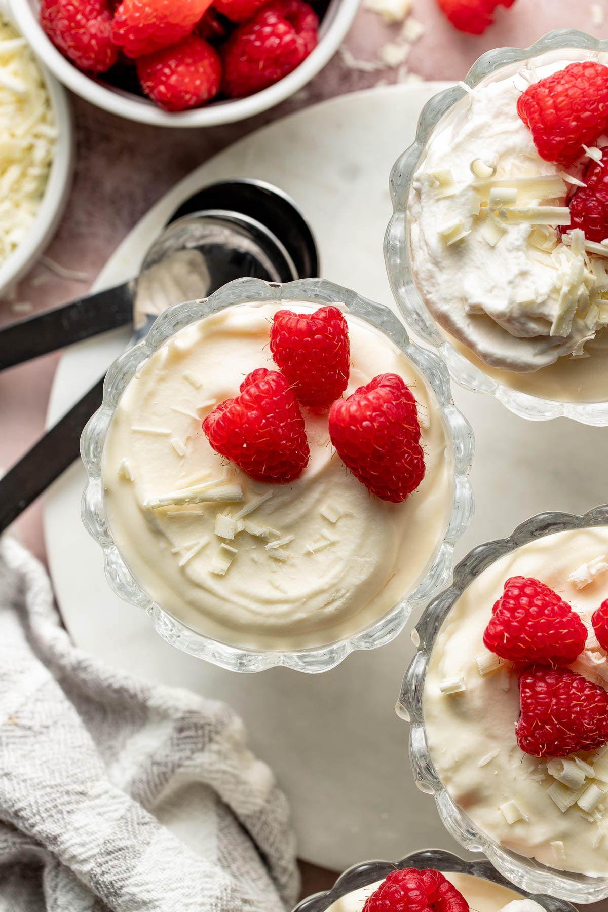 Close overhead view of white chocolate mousse cups garnished with bright raspberries and white chocolate curls. Black spoons, a marble serving board, and bowls of raspberries frame the creamy desserts.