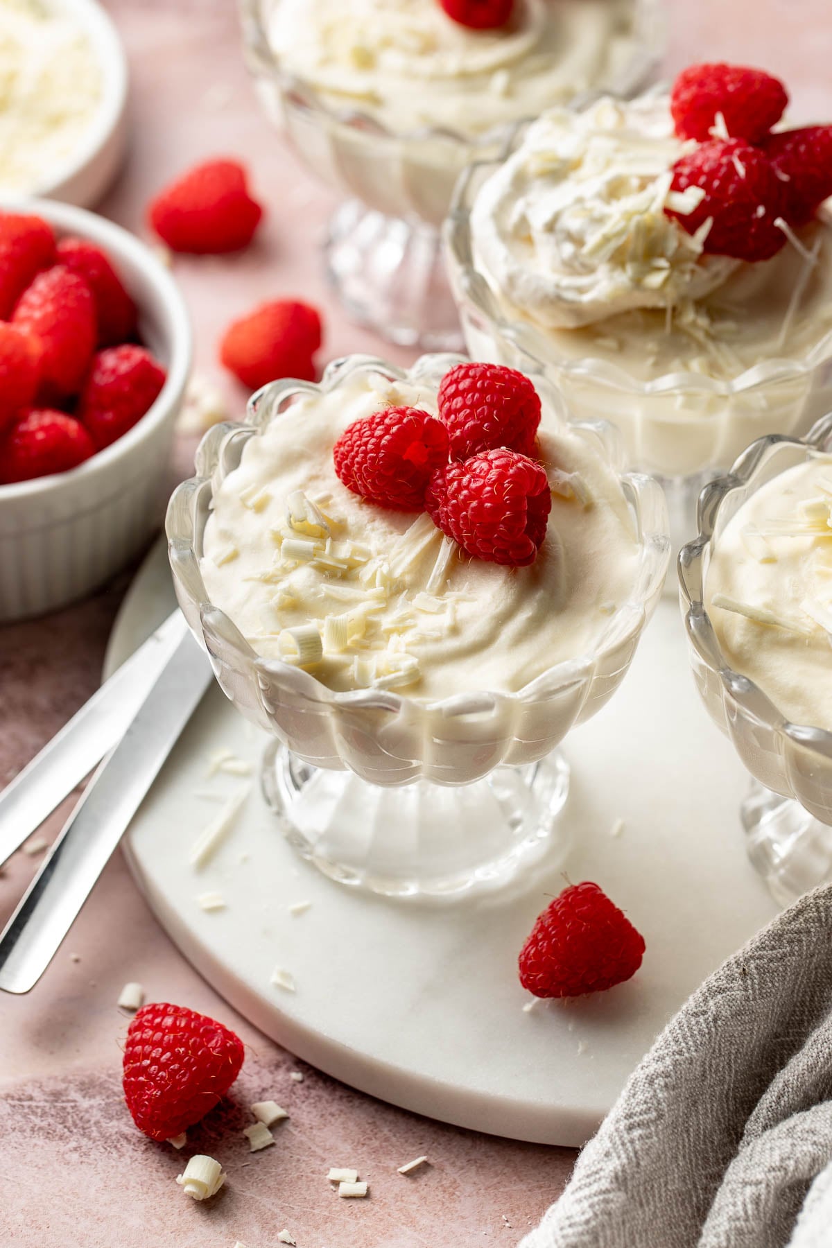Angled close-up of glass dessert cups filled with silky white chocolate mousse and topped with raspberries, whipped cream, and white chocolate shavings. Fresh raspberries and dessert spoons are scattered around the marble board.