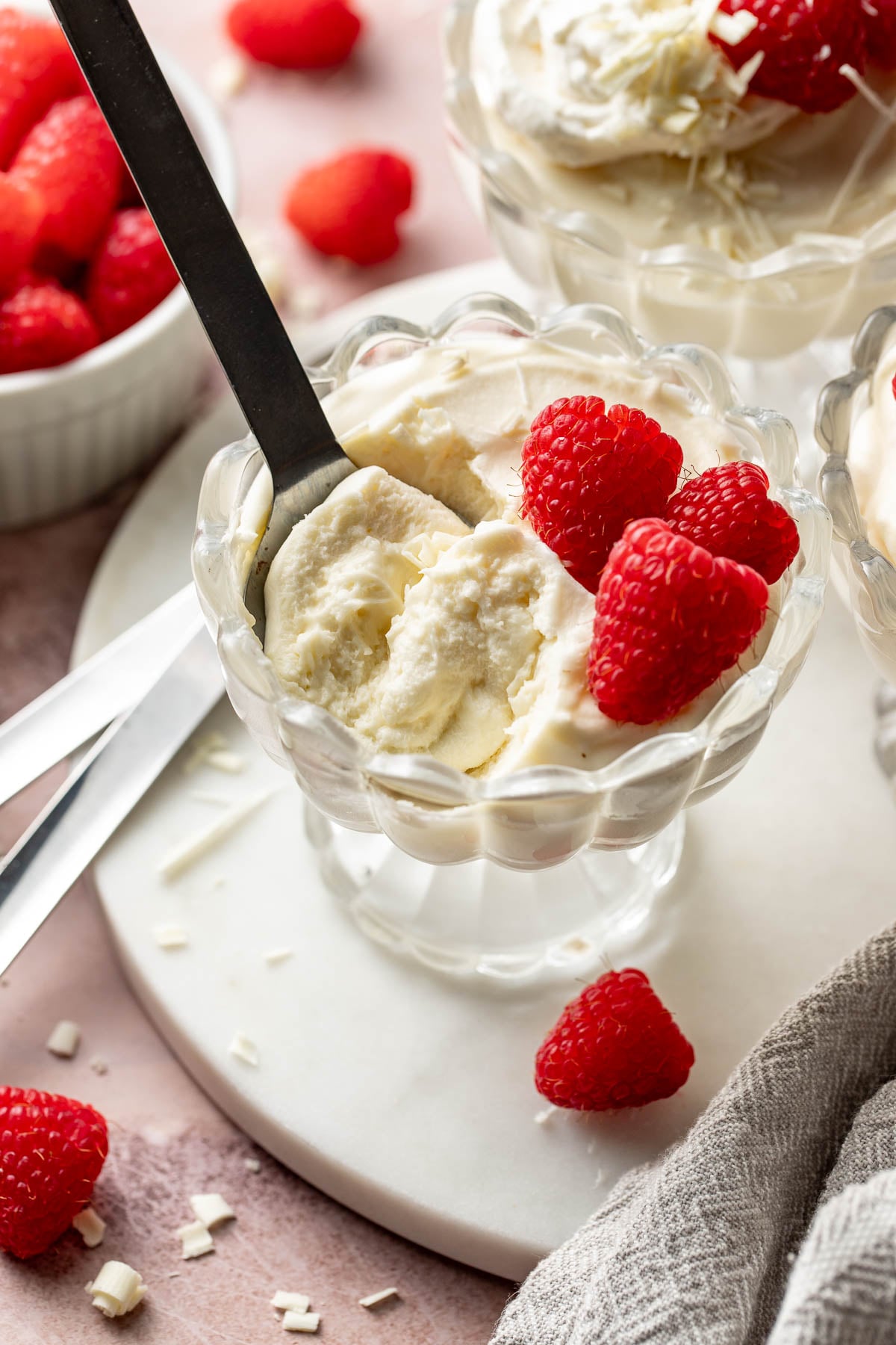 Close-up of a spoon scooping into a glass dish of white chocolate mousse topped with raspberries. The mousse looks light and airy, with white chocolate shavings sprinkled on top.