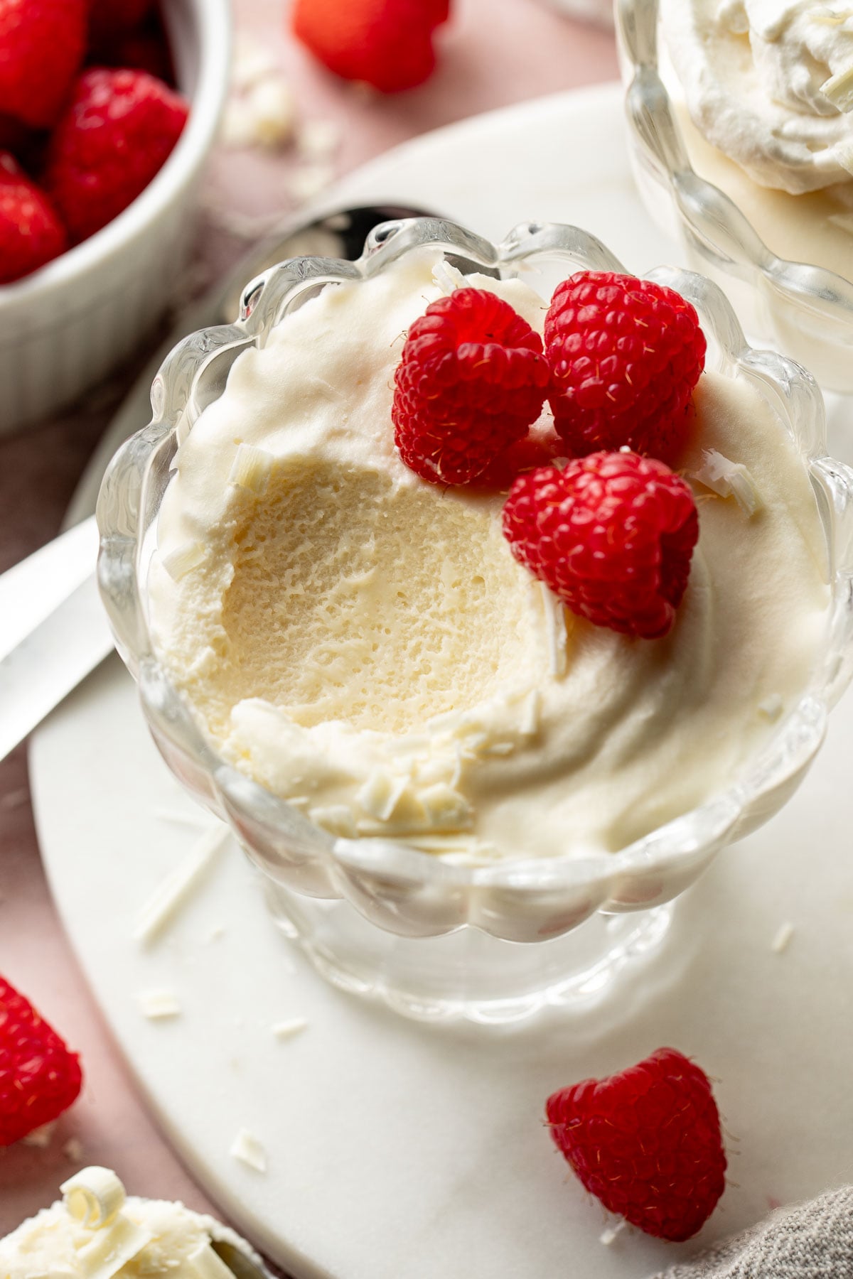 Close-up of a glass dessert cup with a scoop removed, revealing the fluffy, airy texture of the white chocolate mousse. Fresh raspberries and white chocolate curls sit on top.