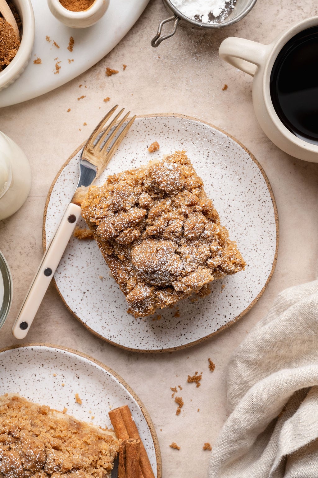 Overhead view of a square slice of cinnamon crumb cake on a speckled plate, dusted with powdered sugar and served with a fork, surrounded by crumbs, cinnamon sticks, and a cup of coffee.