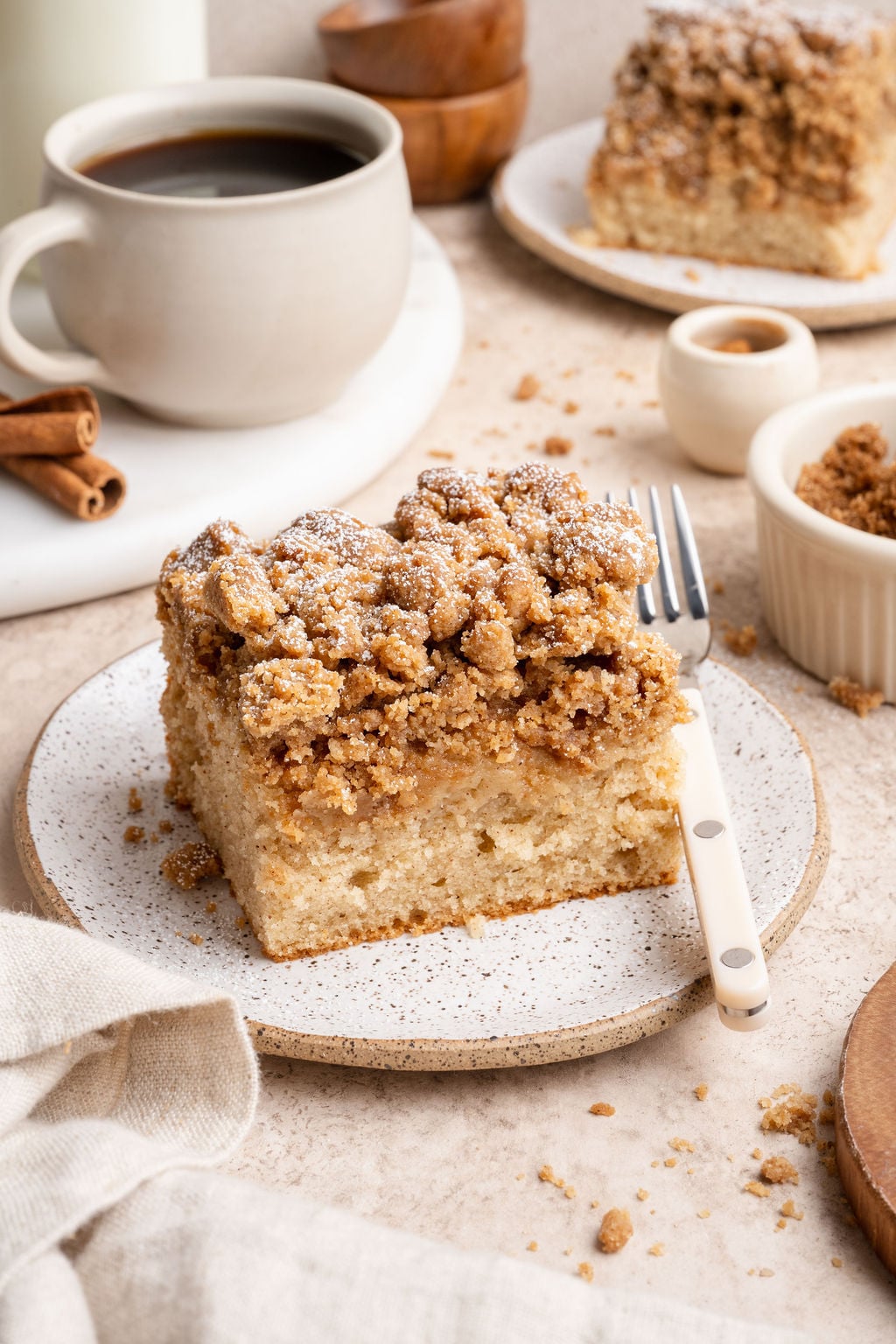 Close-up of a thick slice of cinnamon crumb cake with a soft, tender base and chunky streusel topping, plated with a fork, with a coffee mug and extra crumbs in the background.