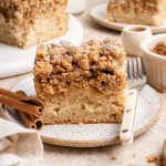 A thick slice of cinnamon crumb cake on a speckled plate, topped with a generous buttery streusel and a light dusting of powdered sugar. Cinnamon sticks and a fork sit beside it, with additional slices of the cake, a cup of coffee, and a small bowl of crumble blurred in the background.