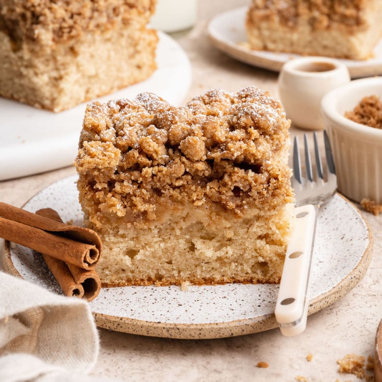 A thick slice of cinnamon crumb cake on a speckled plate, topped with a generous buttery streusel and a light dusting of powdered sugar. Cinnamon sticks and a fork sit beside it, with additional slices of the cake, a cup of coffee, and a small bowl of crumble blurred in the background.