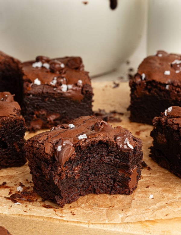 Close-up of a thick brownie with a bite taken out, revealing a dense and fudgy texture with melty chocolate chunks. Set on parchment paper with other brownies nearby.