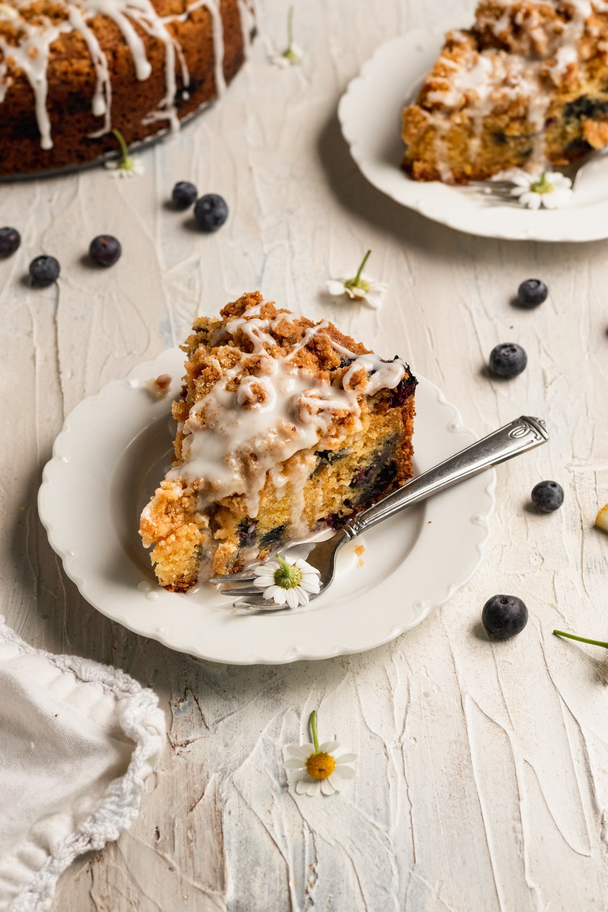 a slice of blueberry coffee cake on a white plate with a lemon glaze on top.