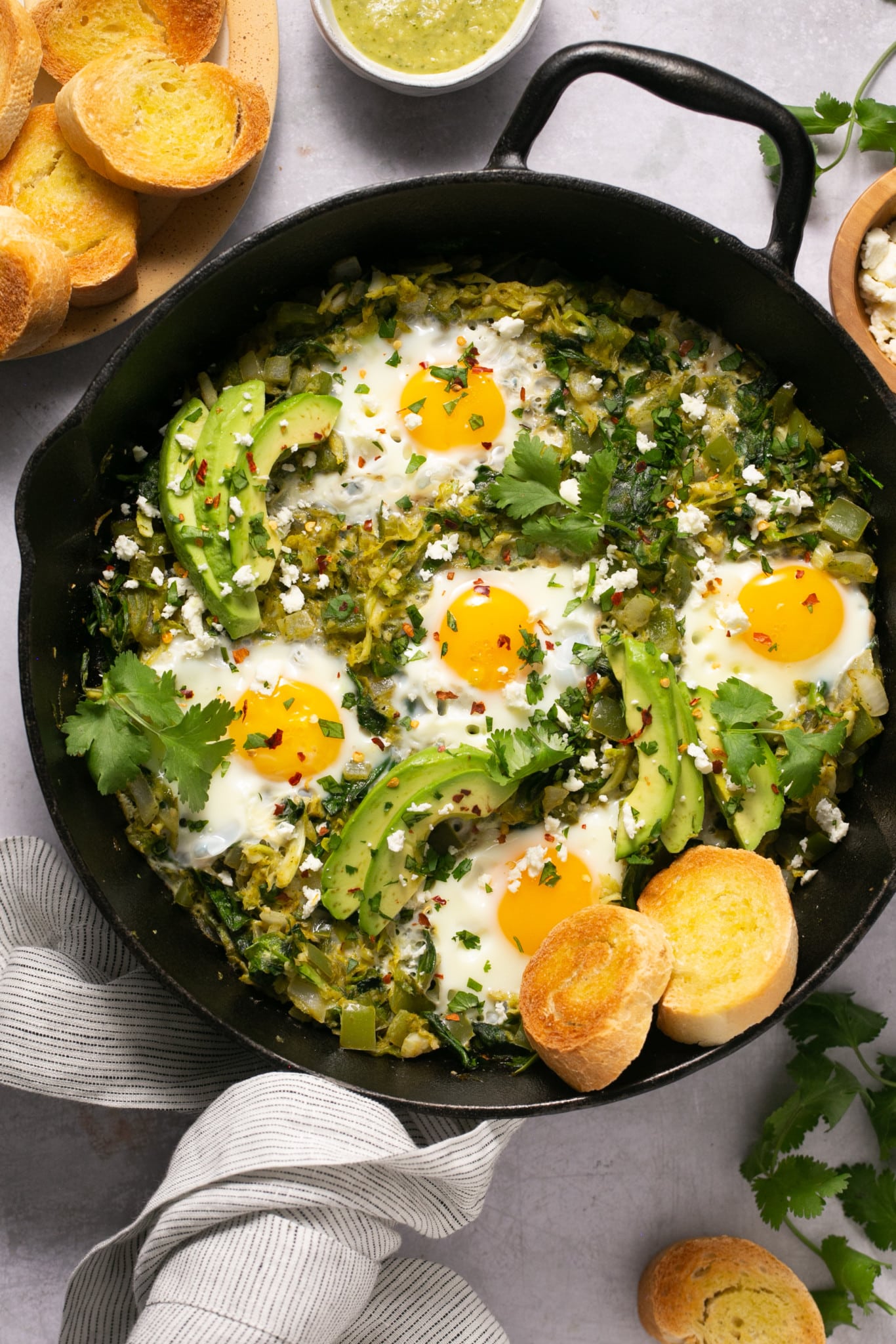 cast iron skillet with eggs, green sauce, avocado, and biscuits