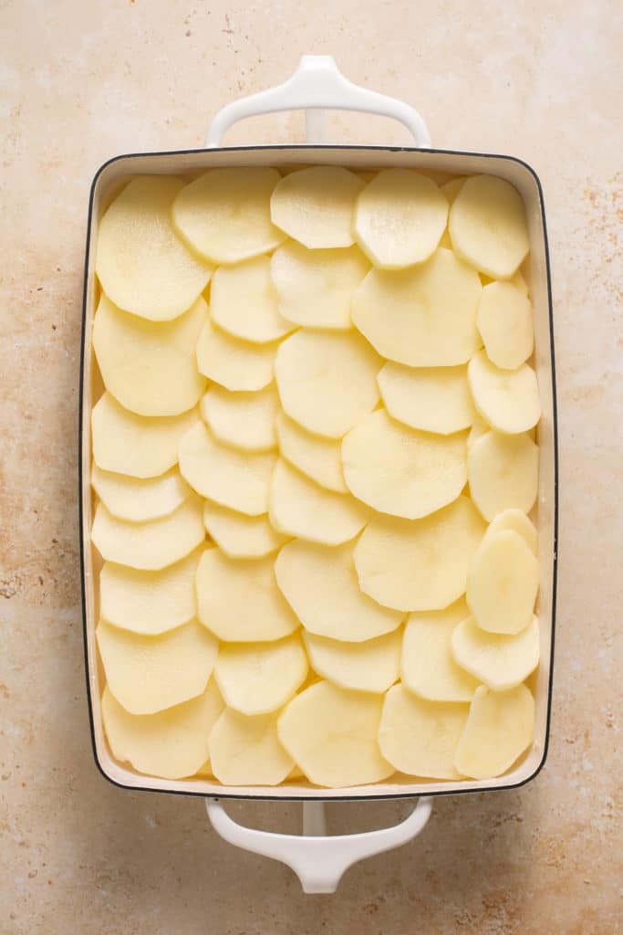 potatoes being layered with cheese sauce in a casserole dish. 