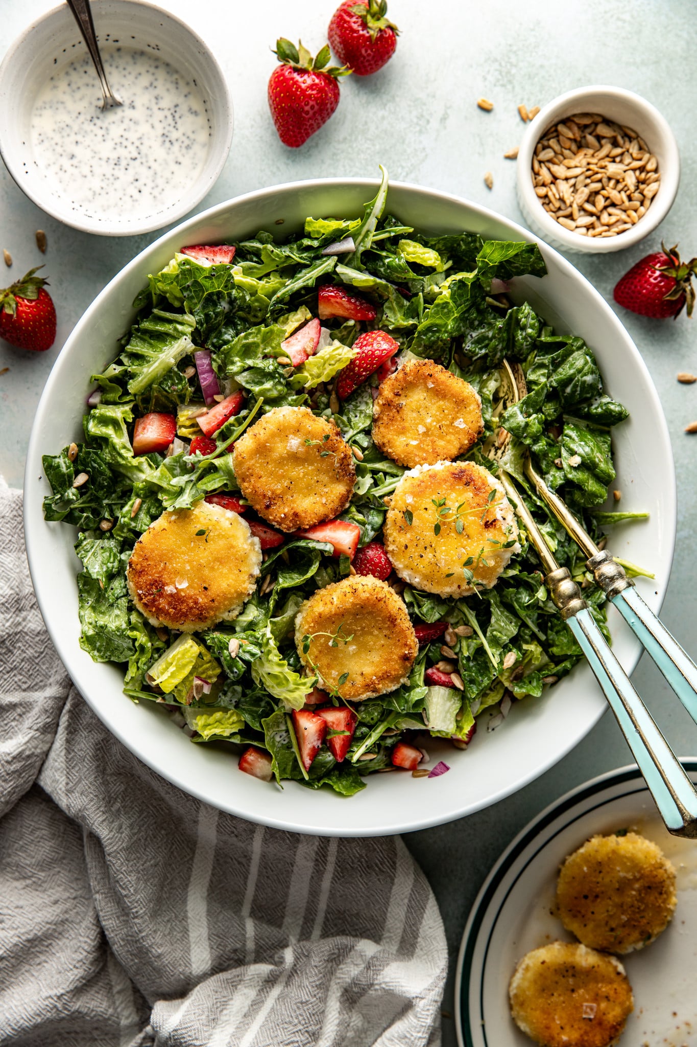 The finished strawberry poppyseed salad topped with golden, pan-fried goat cheese medallions, ready to serve in a white bowl with vintage-style utensils.