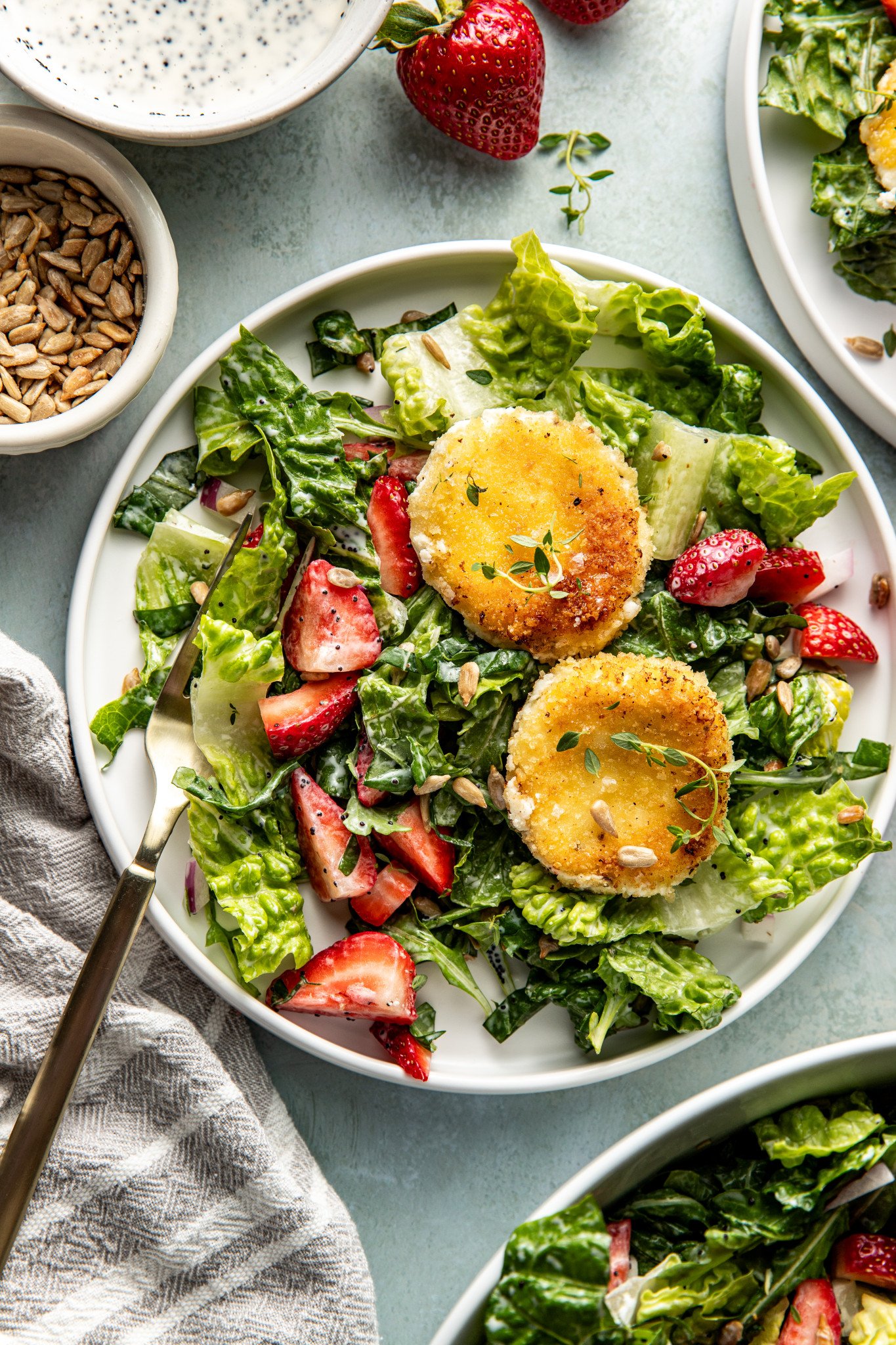 Plated servings of strawberry poppyseed salad on small white plates, each topped with crispy goat cheese rounds and surrounded by bowls of dressing and sunflower seeds.
