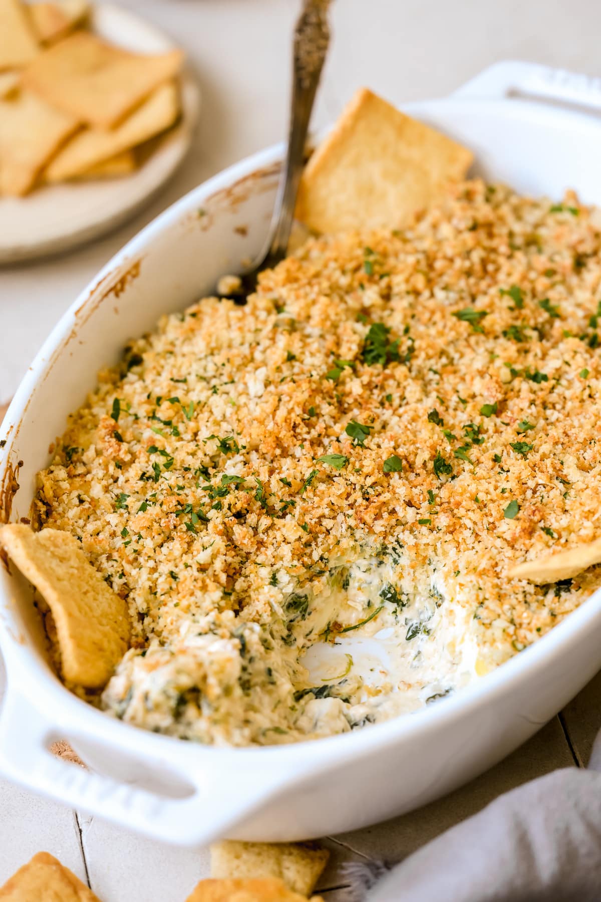artichoke dip in a white baking dish with breadcrumbs and parsley on top. 