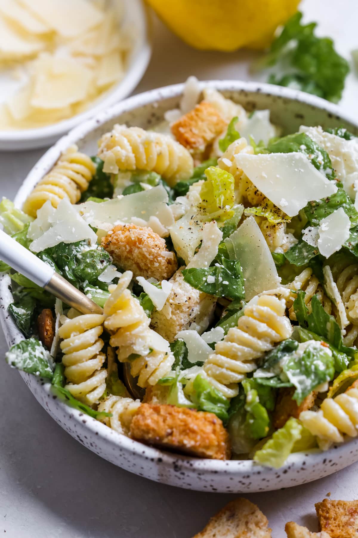 caesar salad on a white speckled plate with parmesan cheese, croutons, black pepper and rotini pasta.