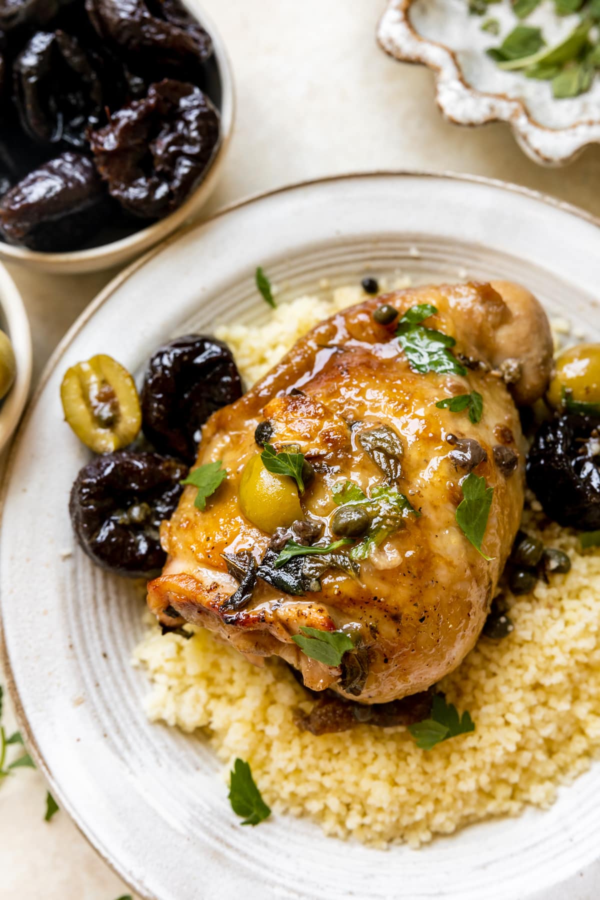 an up close image on a chicken thigh on a white plate with couscous, capers, prunes, olives and parsley garnished on top. 