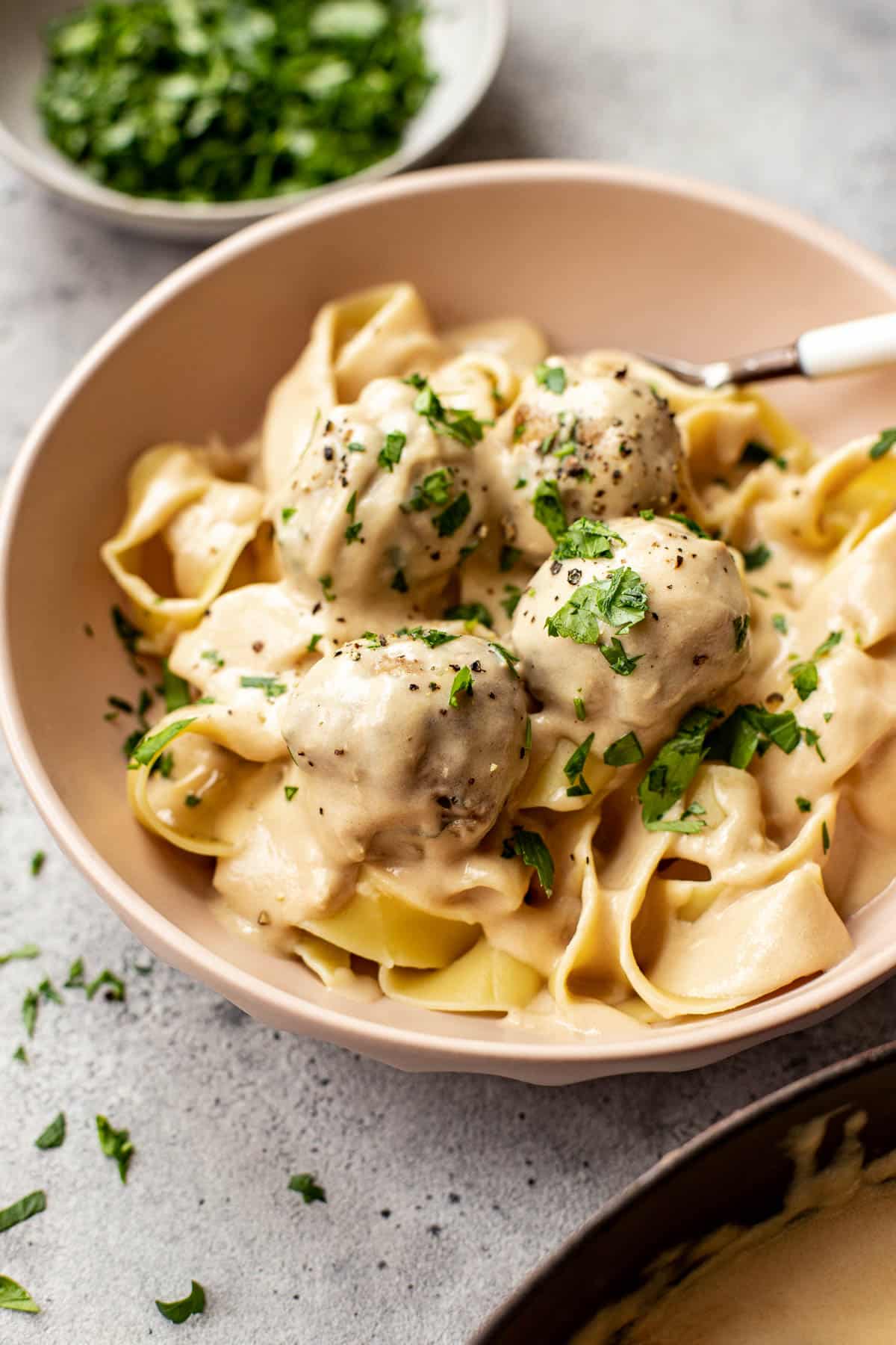 pasta and meatballs in a light pink bowl with parsley and black pepper on top. 