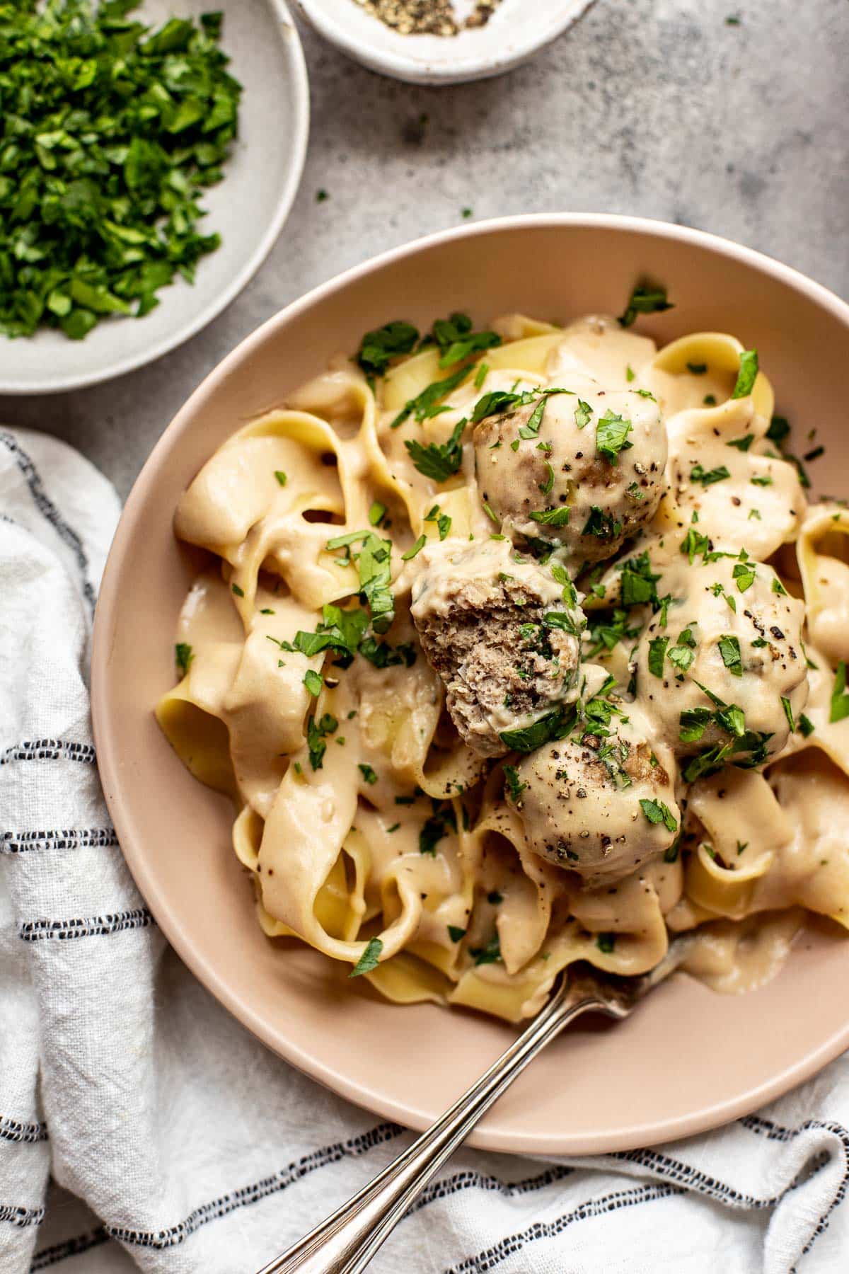 a bite taken out of a meatball in a pink bowl with pasta and parsley garnished on top. 