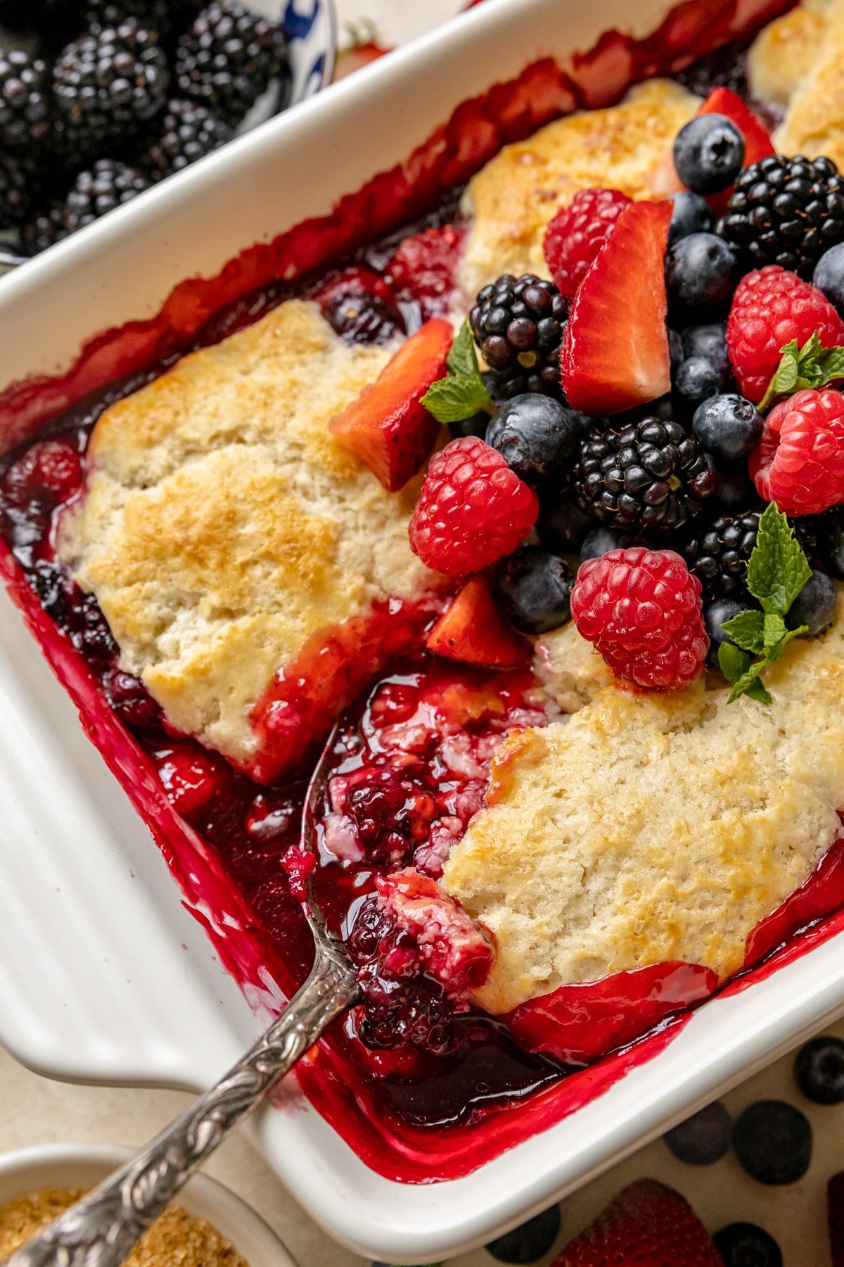 a spoon scooped into a berry cobbler with fresh berries and mint on top.