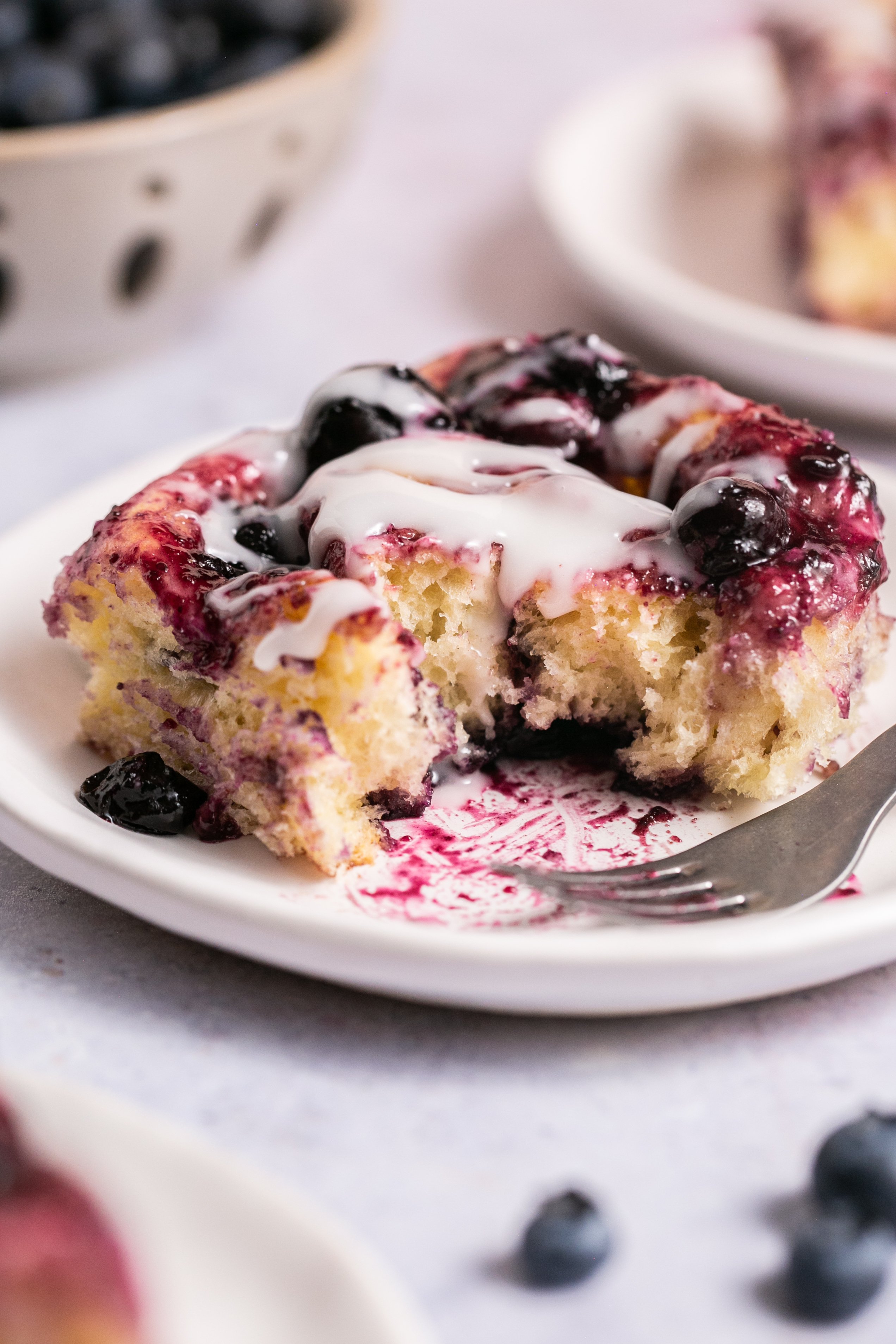 Cinnamon roll on a white plate covered in a glaze and blueberry sauce. 