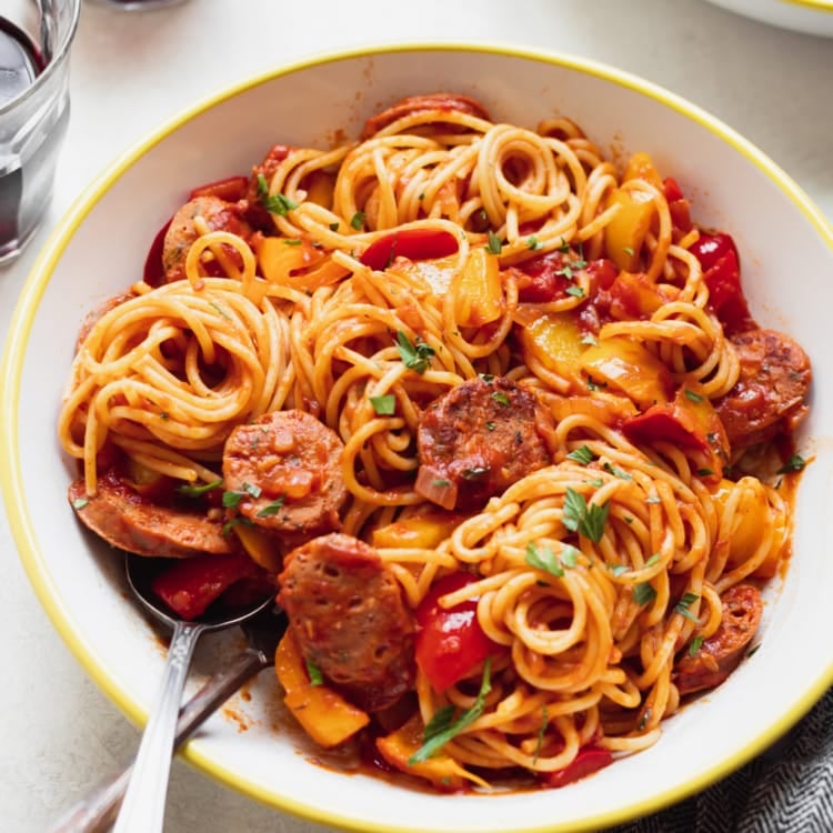 spaghetti noodles in a white bowl with a yellow rim, with red sauce, peppers, and sausages