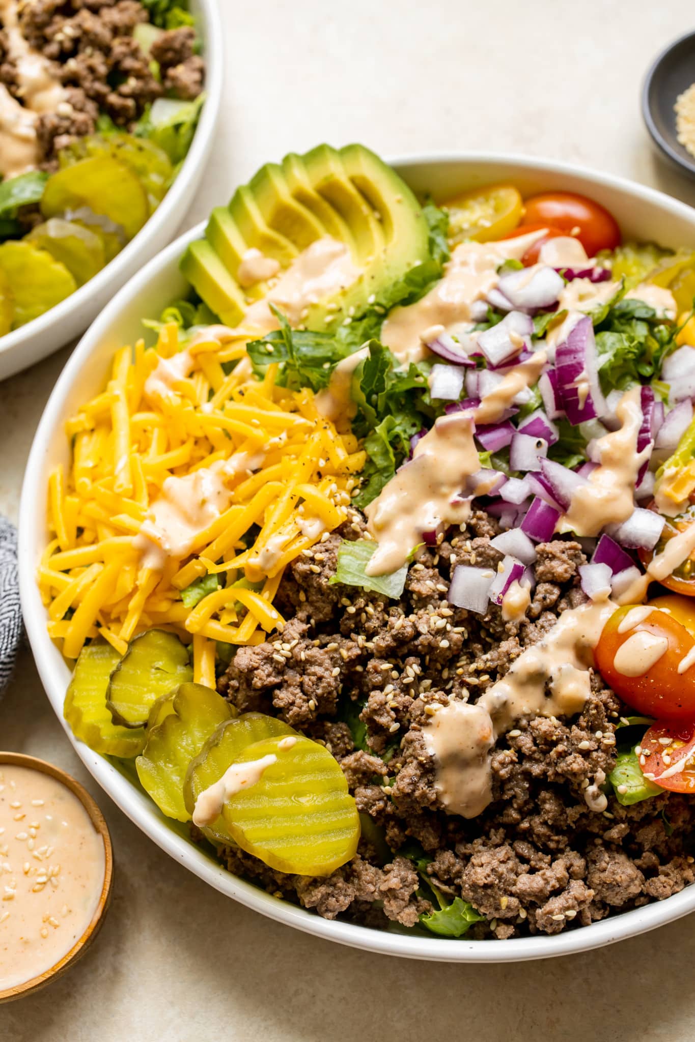 romaine lettuce in a white bowl topped with burger toppings like ground beef, cheese, avocado, red onion, pickles, tomatoes and a special burger sauce. 