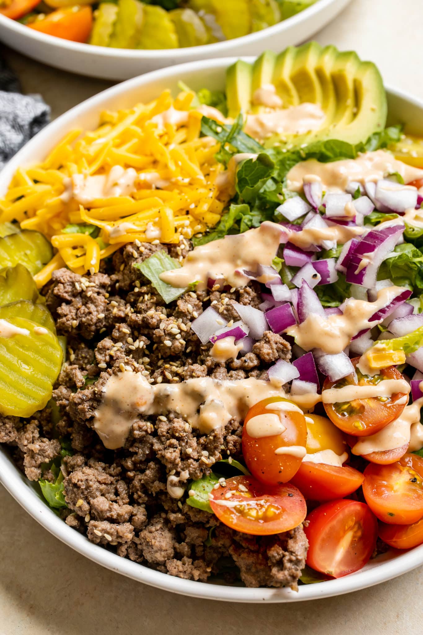 romaine lettuce in a white bowl topped with burger toppings like ground beef, cheese, avocado, red onion, pickles, tomatoes and a special burger sauce. 