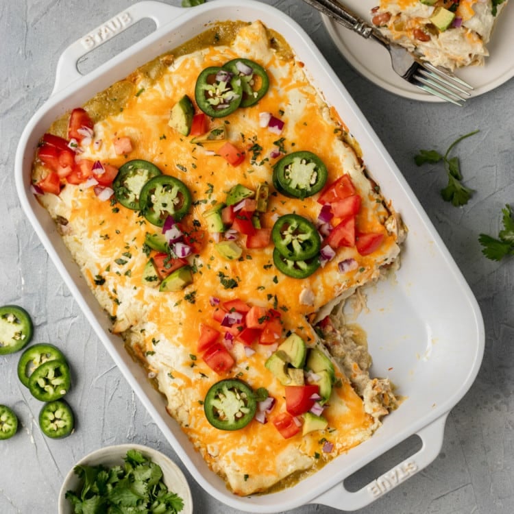 white casserole dish with tortillas layered with chicken topped with shredded cheese, jalapeno, tomatoes, avocado