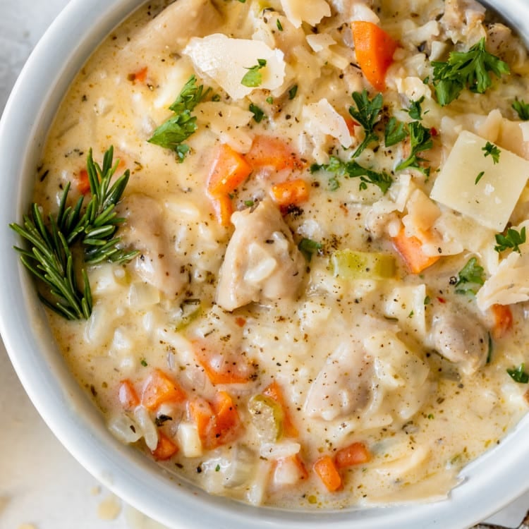 white bowl filled with a creamy soup with rice, carrots, onions, chicken, celery, parmesan cheese, and green herbs
