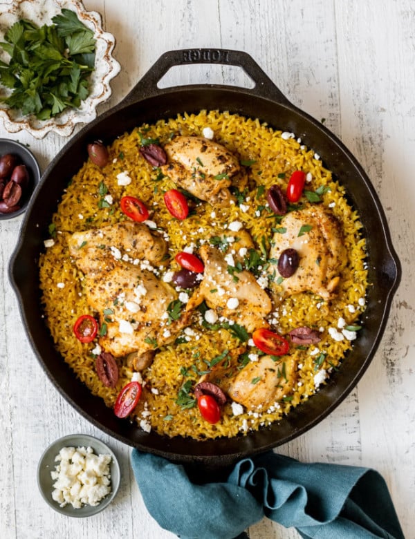 black cast iron skillet with yellow rice, chicken thighs, cherry tomatoes, feta cheese, kalamata olives, and green spices