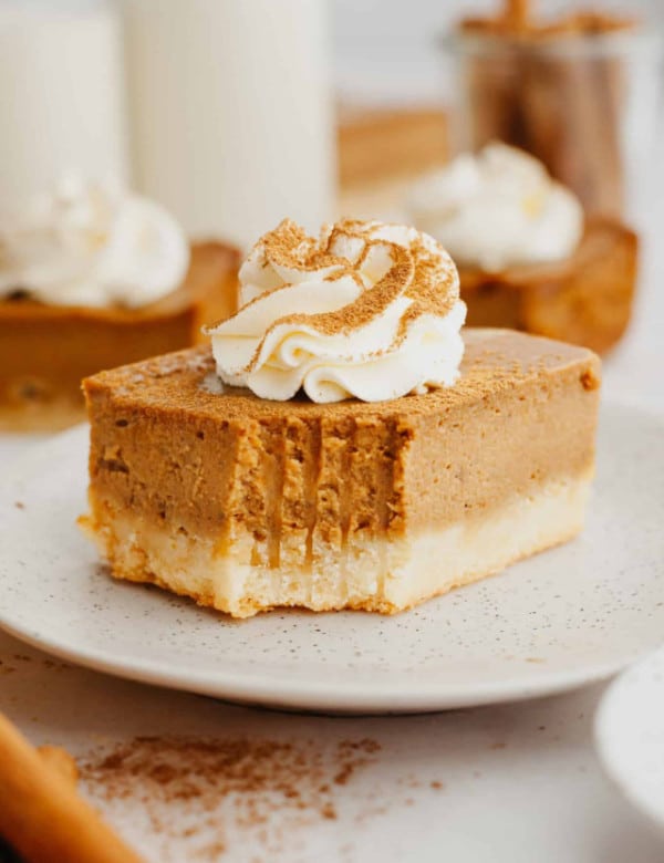 one slice of pumpkin pie bars with a bite missing on a tan plate and topped with whipped cream and a dust of spices