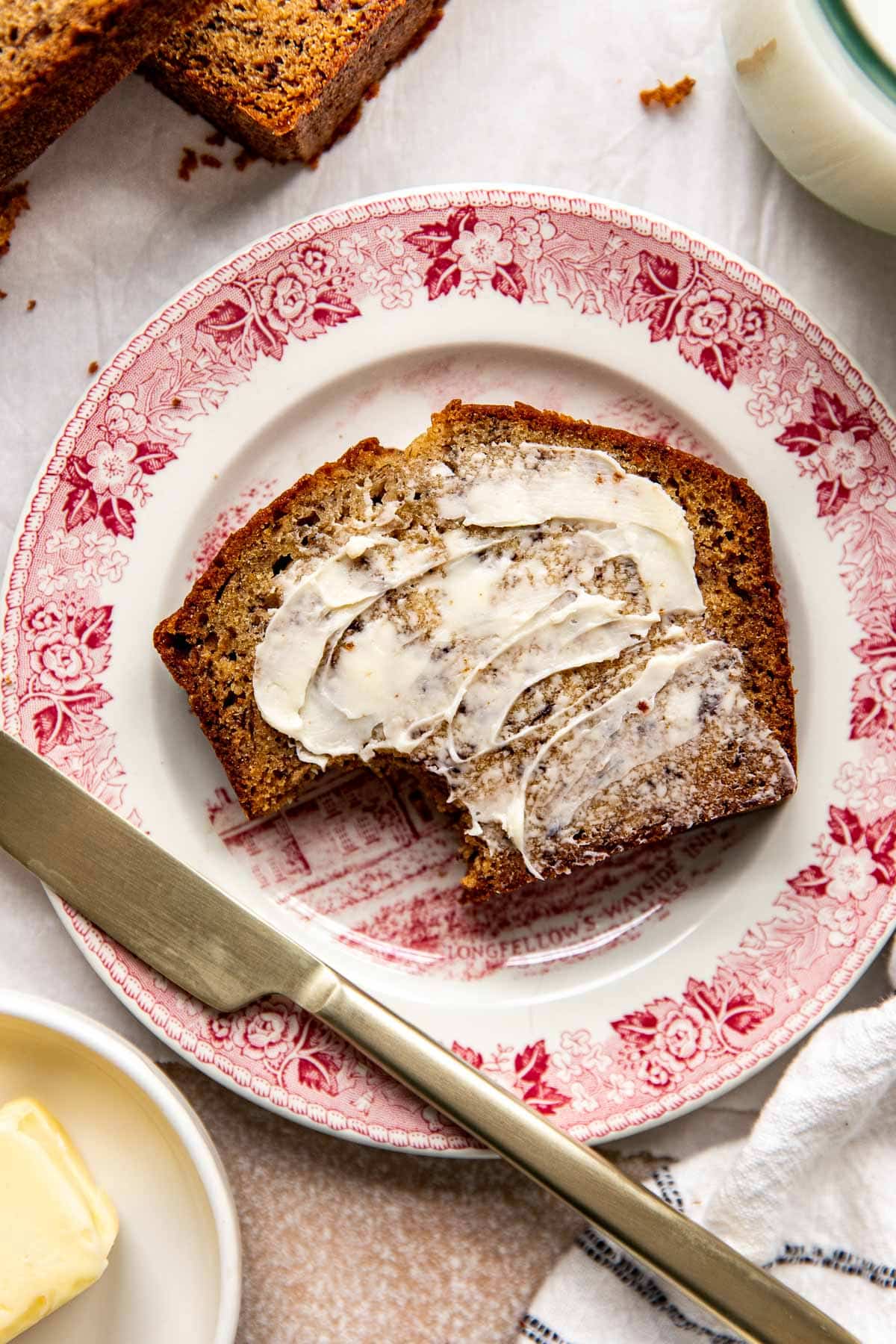 a slice on banana bread with butter on top on a red and white plate with a gold knife. 