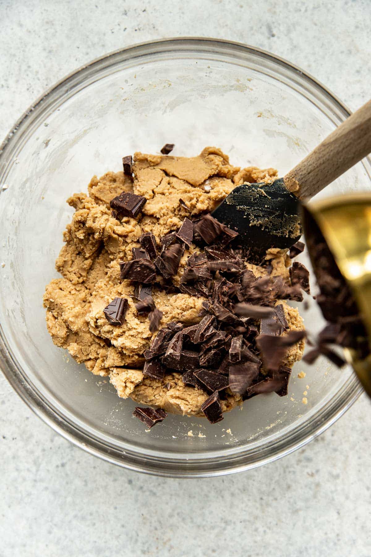 chocolate chunks being poured into the cookie dough in a large glass bowl. 