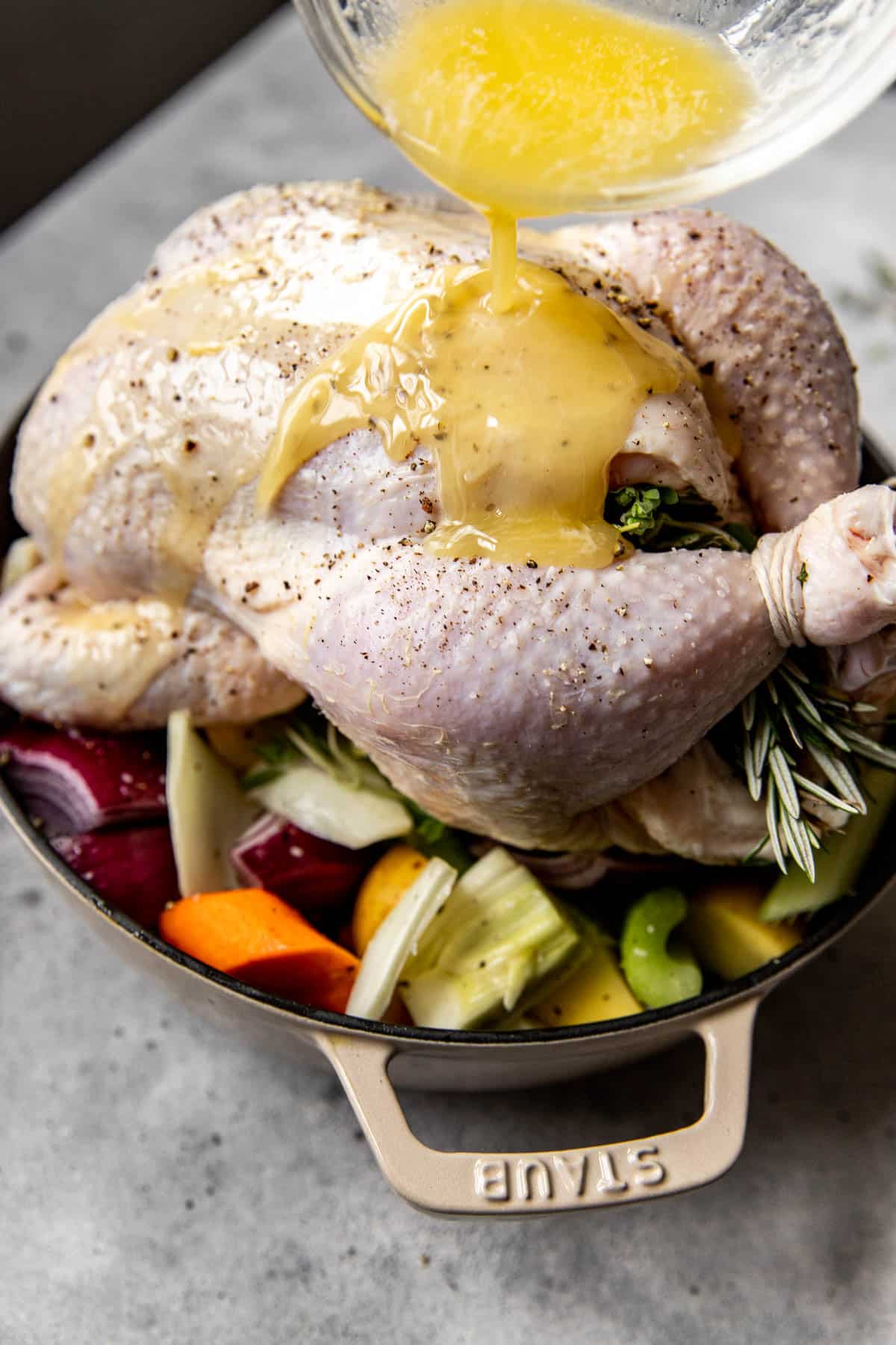 melted butter being poured on a chicken in a roasting pan on top of a variety of vegetables. 