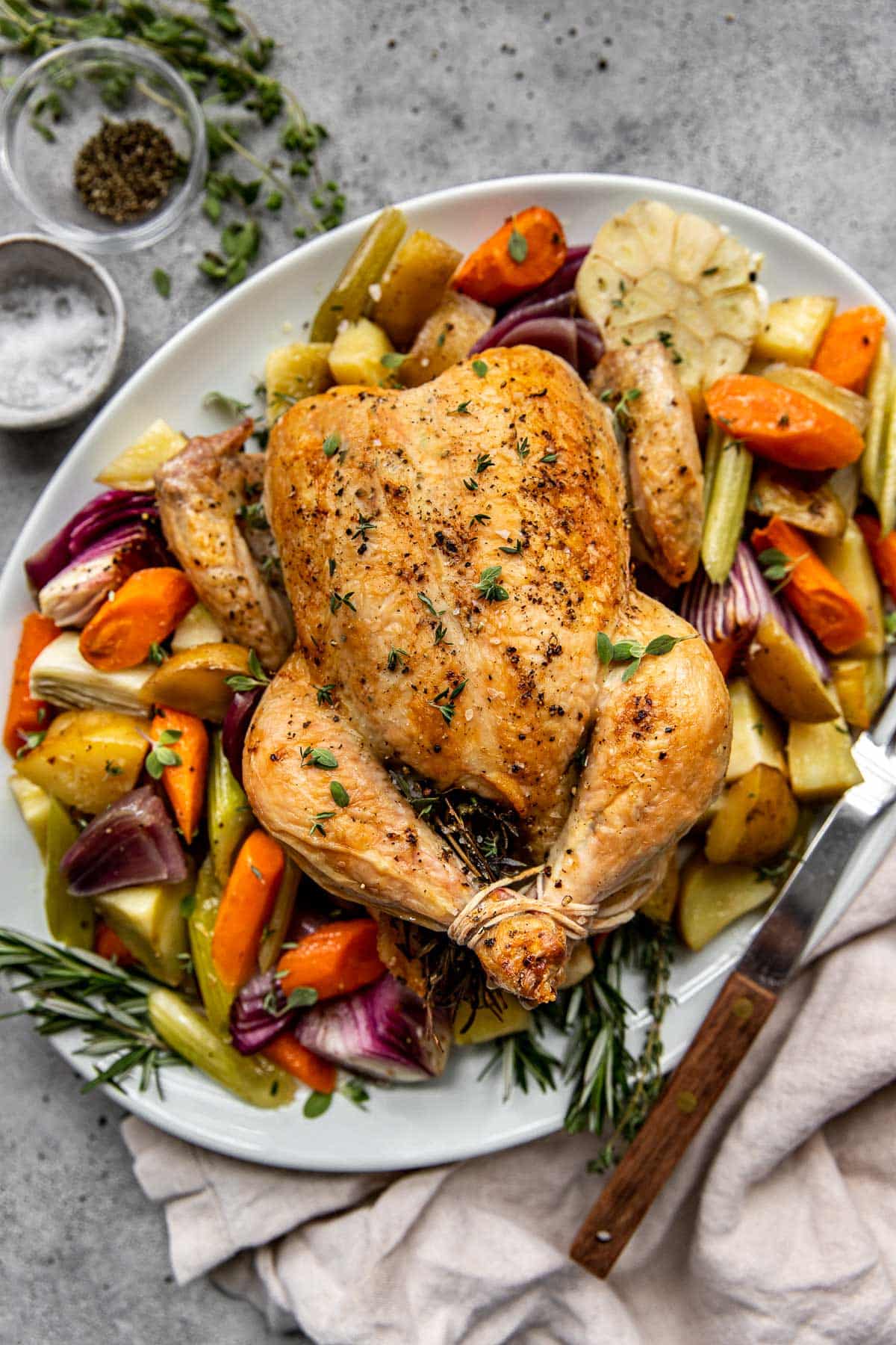 a roasted chicken on a white serving platter with colorful vegetables and fresh herbs. 