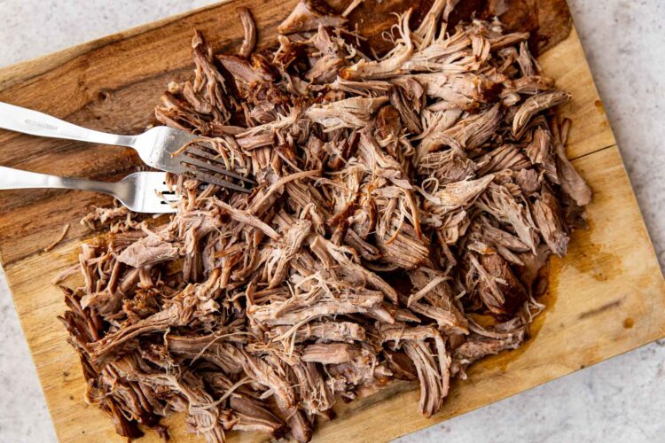 two forks shredding pulled pork on a wooden cutting board.