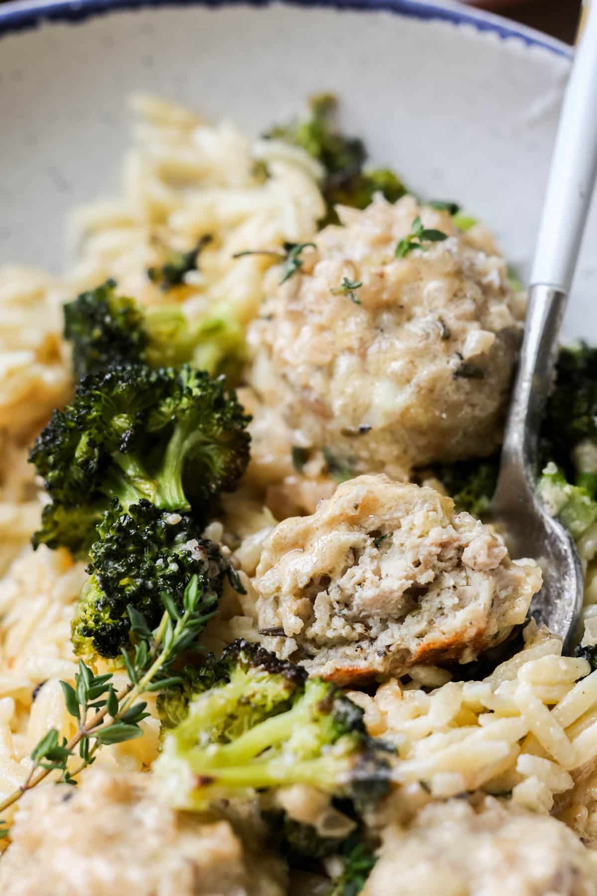 a fork taking a bite out of a chicken meatball with broccoli and orzo. 
