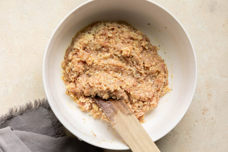 Ground chicken meatball mixing in a white bowl with a wooden spoon.