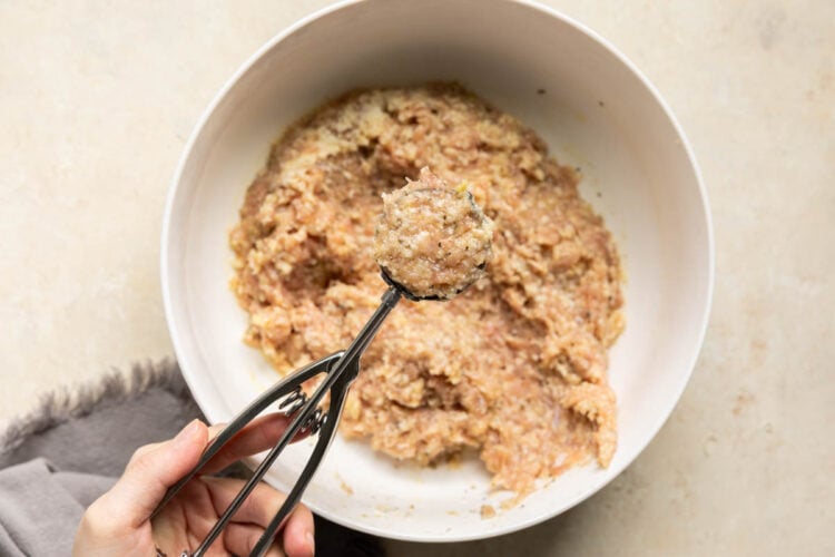 ground chicken meatball mixture in a white bowl with a cookie scoop scooping some of the mixture out.