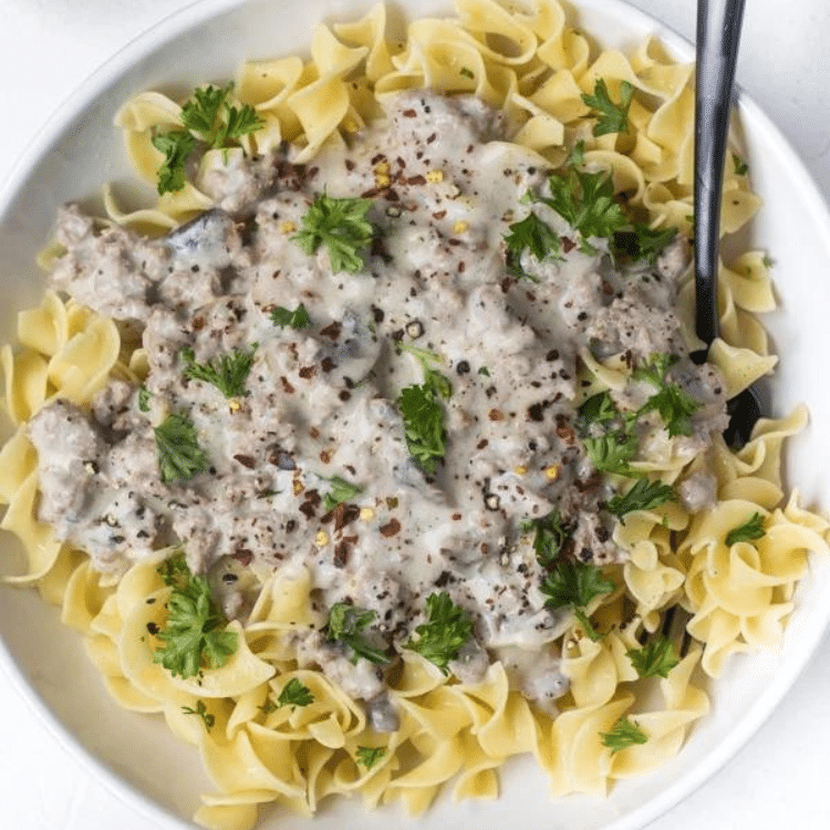 white bowl with egg noodles, stroganoff sauce, and green herbs