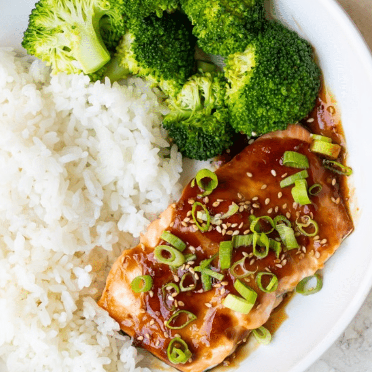 white bowl with white rice, broccoli, and a salmon fillet topped with sauce, green onions, and sesame seeds