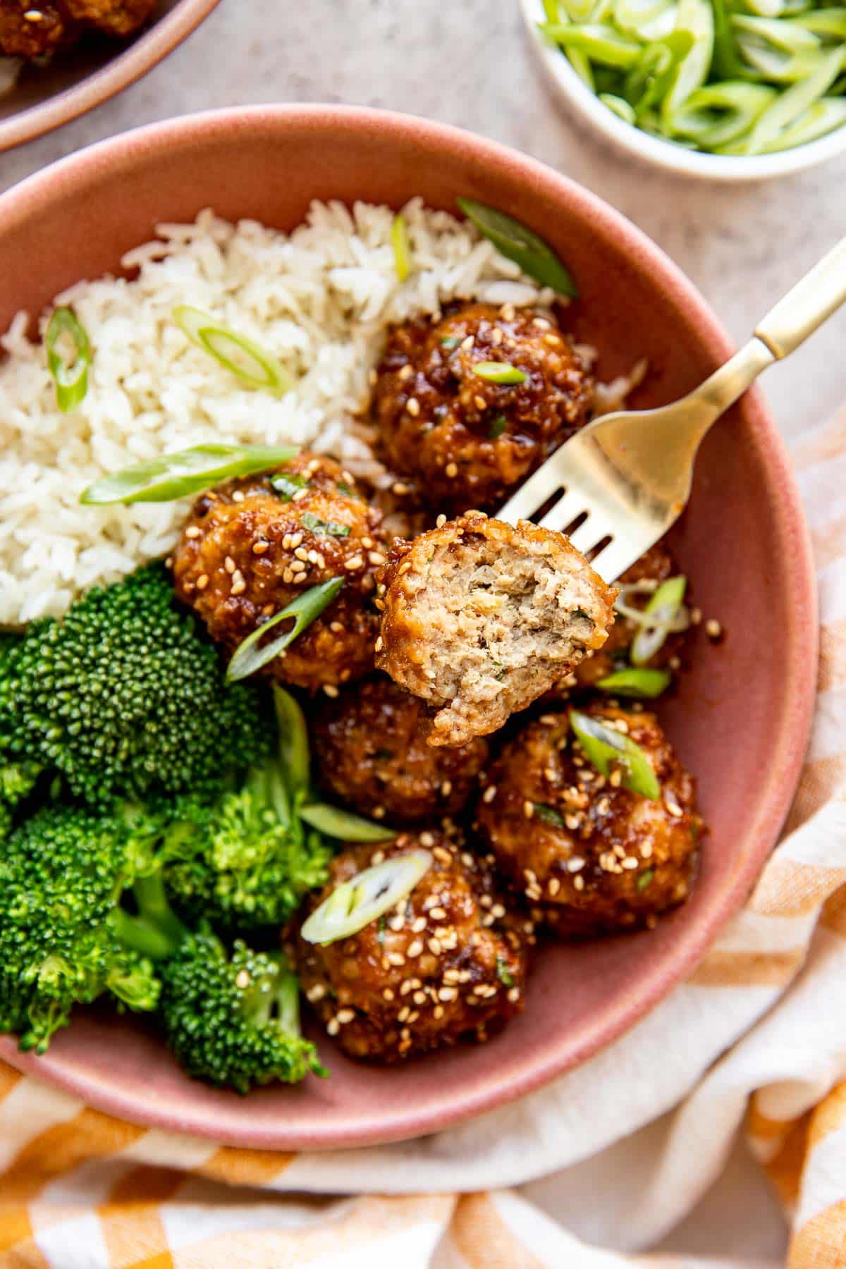 a gold fork that has taken a bite out of a sriracha meatball in a bowl with broccoli and rice. 